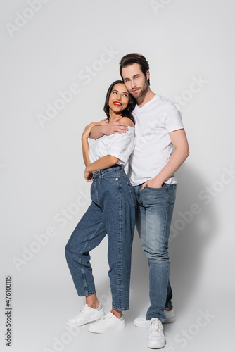full length view of young man embracing brunette woman while standing with hand in pocket of jeans on grey.