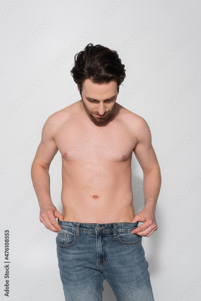 young shirtless man touching jeans while standing on grey.