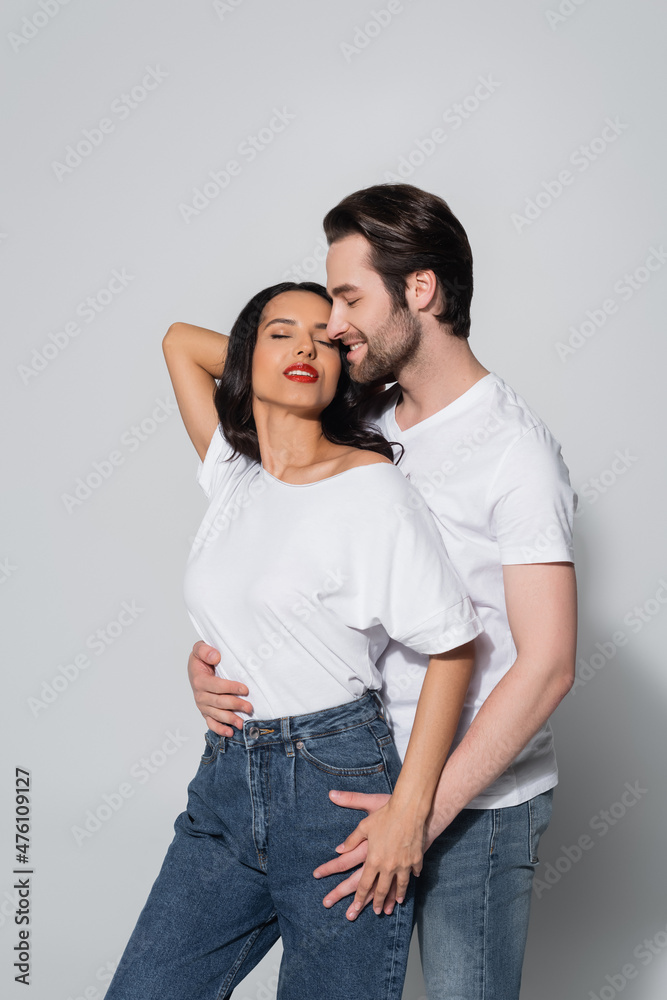 smiling man hugging seductive woman standing with hand behind head and closed eyes on grey.