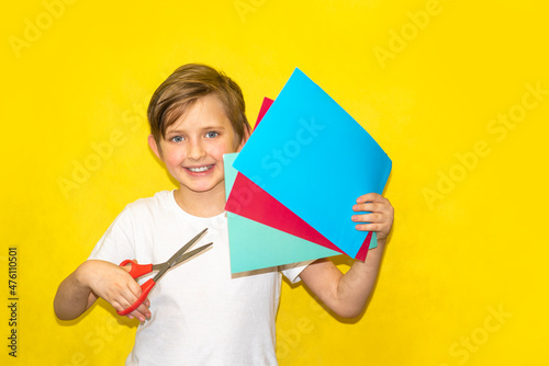 a happy boy in a white shirt holds scissors in one hand and colorful paper in the other hand on a yellow background