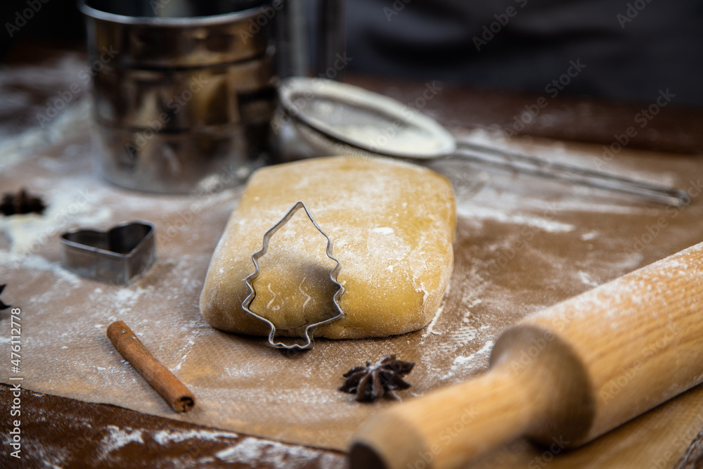 A cookie cutter in the form of a Christmas tree is leaning on the dough, cinnamon and cloves are lying next to it, flour, other cookie cutters and a rolling pin for dough are visible in the background A cookie cutter in the form of a Christmas tree is leaning on the dough, cinnamon and cloves are lying next to it, flour, other cookie cutters and a rolling pin for dough are visible in the background