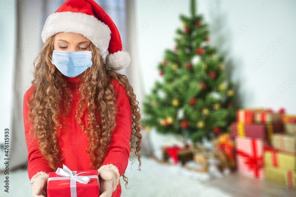 Portrait of a beautiful girl in medical mask and santa hat with a present in hands, standing in front of decorated christmas tree at home