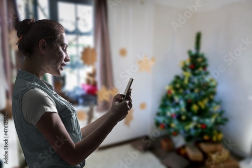 Happy woman using phone, celebrating success, sitting on Christmas tree background at home.