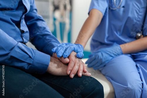 Close up of nurse holding hands of elder patient for support in medical office. Healthcare specialist with gloves giving assistance to man after osteopathy examination and results