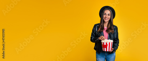 Banner photo of stylish young beautiful hipster woman in black jacket and hat with a bucket of popcorn while she is posing isolated on yellow background