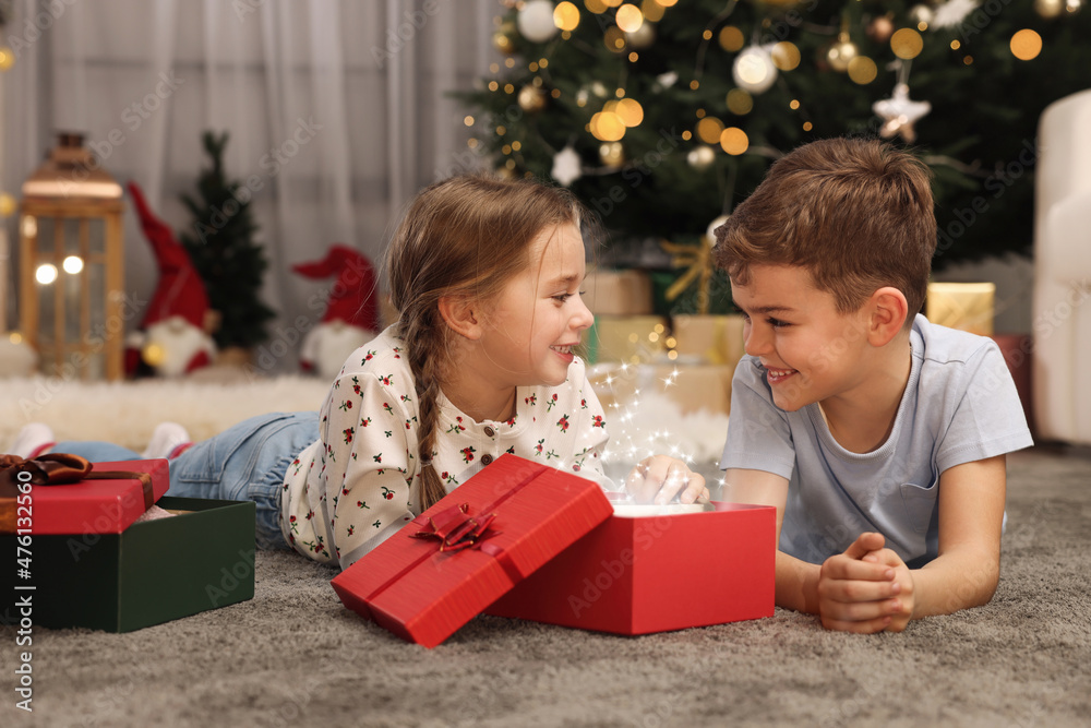 Cute little children opening gift box near Christmas tree at home