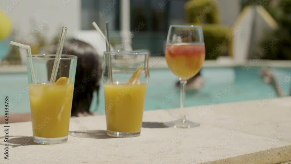 Orange refreshing drinks on swimming pool side. Close-up shot of glasses of alcohol and non alcohol cocktails with straws on background of water and family playing ball in pool. Summer party concept Orange refreshing drinks on swimming pool side. Close-up shot of glasses of alcohol and non alcohol cocktails with straws on background of water and family playing ball in pool. Summer party concept