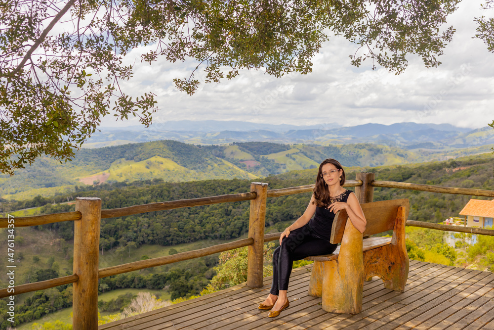 Jovem Mulher vestida de preto sentada em um banco com uma bela paisagem de montanhas e natureza ao fundo. 