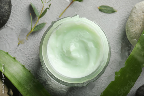 Flat lay of body cream in jar with aloe and eucalyptus on light grey stone table, view through wet glass