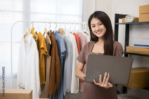 Young beautiful asian woman business owner at fashion store holding laptop check stock on rails clothing. Asian female entrepreneur working in clothes shop.