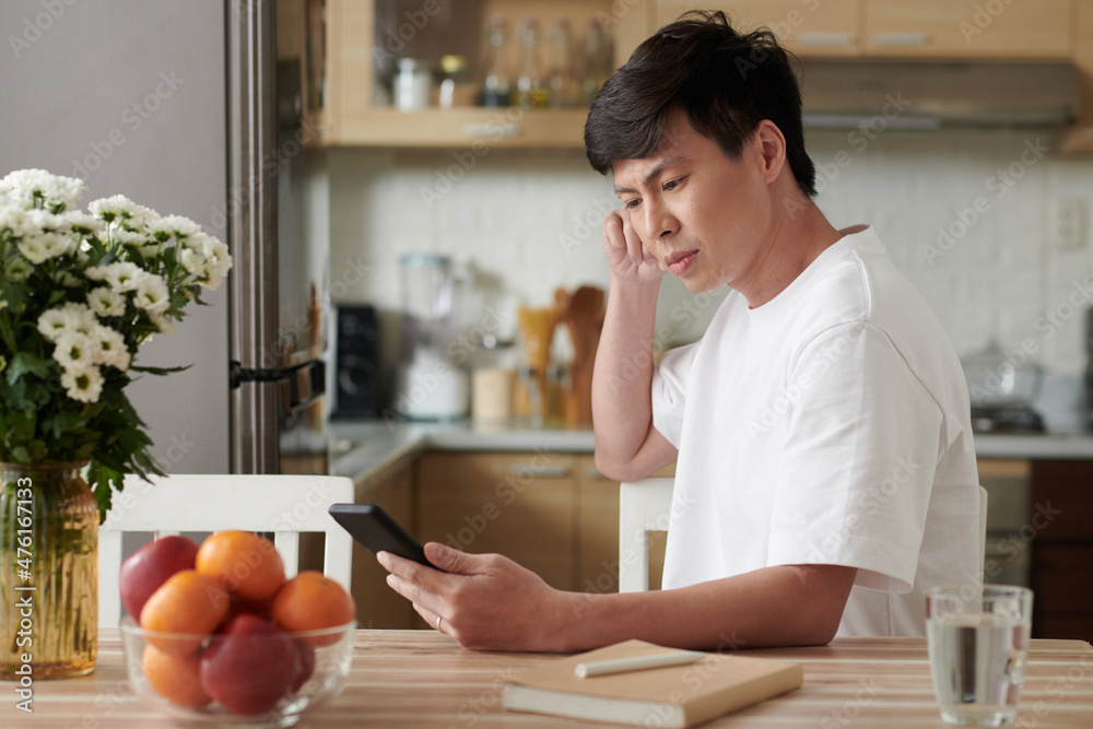 Frowning serious man sitting at table in kitchen and reading article on smartphone screen Frowning serious man sitting at table in kitchen and reading article on smartphone screen