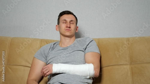 Young man in grey t-shirt touches injured forearm spoke bone in gypsum bandage and sighs tiredly sitting on soft sofa in living room. photo