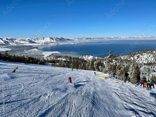 Scenic view of skiers and snowboarders on the snow covered slopes at a ski resort on a bluebird winter day, with Lake Tahoe in the distance