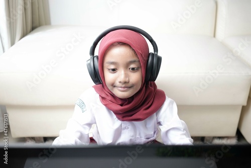 Asian moslem little girl wearing red hijab doing something on her laptop with headphone, for school activity.