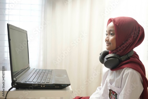 Asian moslem little girl wearing red hijab doing something on her laptop with headphone, for school activity.