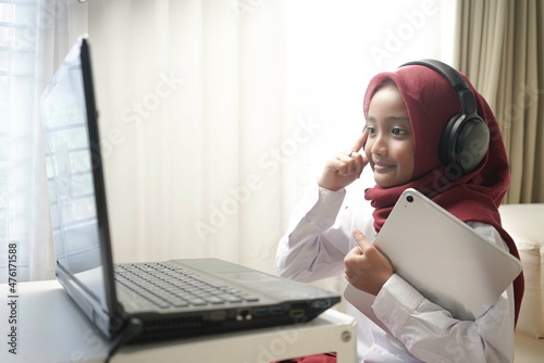 a cute little girl wearing red hijab doing something on her tablet computer and laptop. and wearing headphone