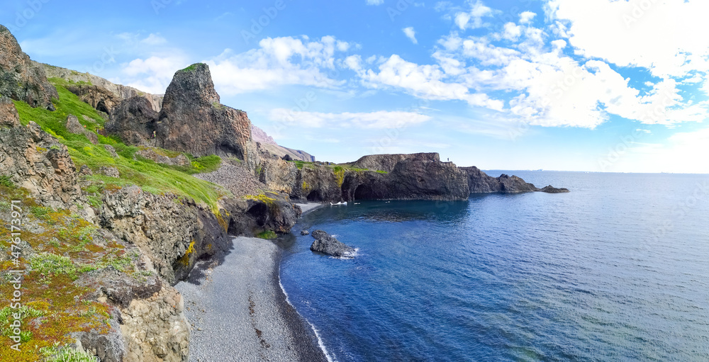 Scenic view of rocky coast with green grass and blue ocean; Person standing on the rock  in a distance with blue ocean,  black sand beach and green coastline with rocks and cliffs and blue cloudy sky