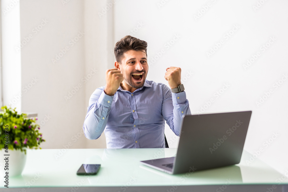 Young businessman in suit looking at laptop excited by good news online, lucky successful winner man sitting at office desk raising hand in yes gesture celebrating business success win result Young businessman in suit looking at laptop excited by good news online, lucky successful winner man sitting at office desk raising hand in yes gesture celebrating business success win result