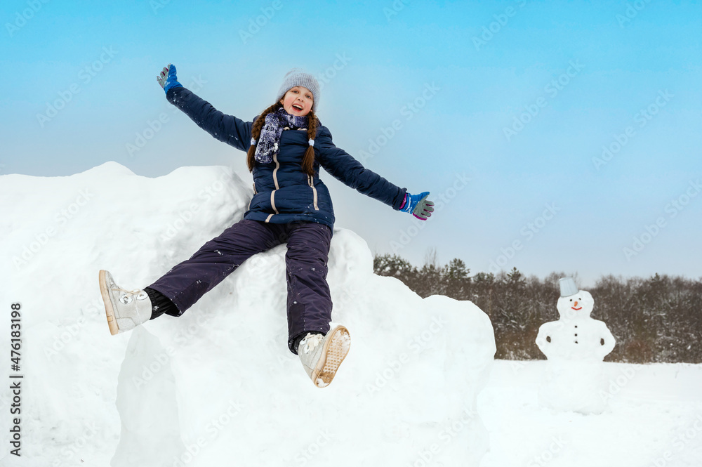 teenage girl playing with snow on a winter day, sitting on a snow fortress, a snowman in the background. Winter outdoor activities teenage girl playing with snow on a winter day, sitting on a snow fortress, a snowman in the background. Winter outdoor activities