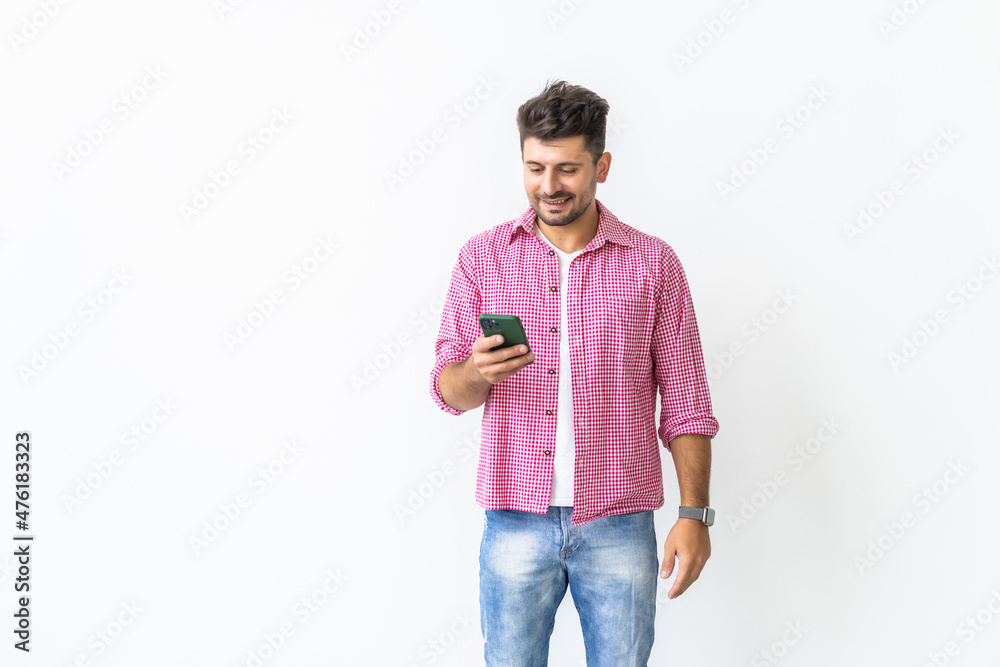 Fototapeta premium Confident young handsome man in jeans shirt holding smart phone and looking at it with smile while standing against white background