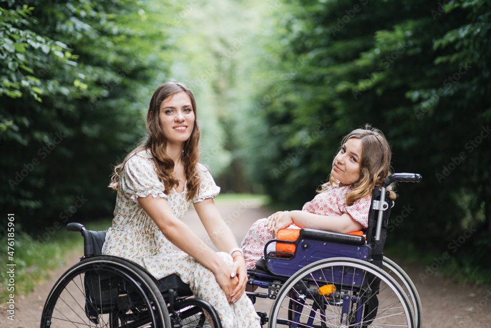 Obraz premium Portrait of young caucasian ladies in wheelchairs sitting in front of each other at summer park and looking at camera. Concept of people with disability and lifestyles.