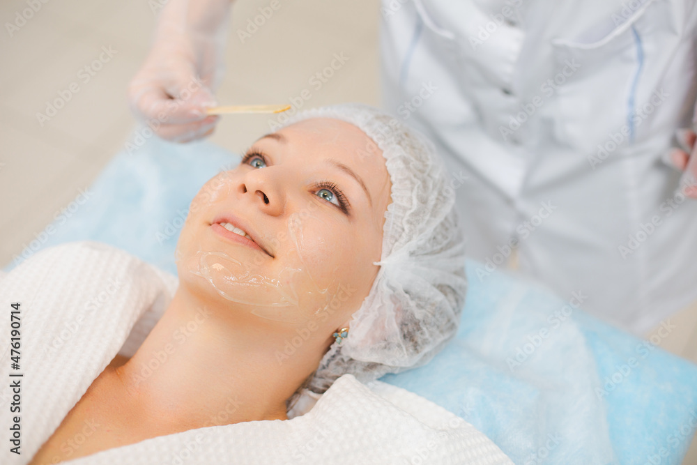 Hands of cosmetology specialist applying facial mask using stick, making skin hydrated and face glowing. Attractive woman relaxing smiling and enjoying spa procedures