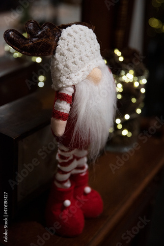 Ozdoba świąteczna, skrzat, krasnal, elf z białą brodą, czapką i porożem renifera. Ubrany w czerwono-białe ubranko w paski. W tle światełka choinkowe