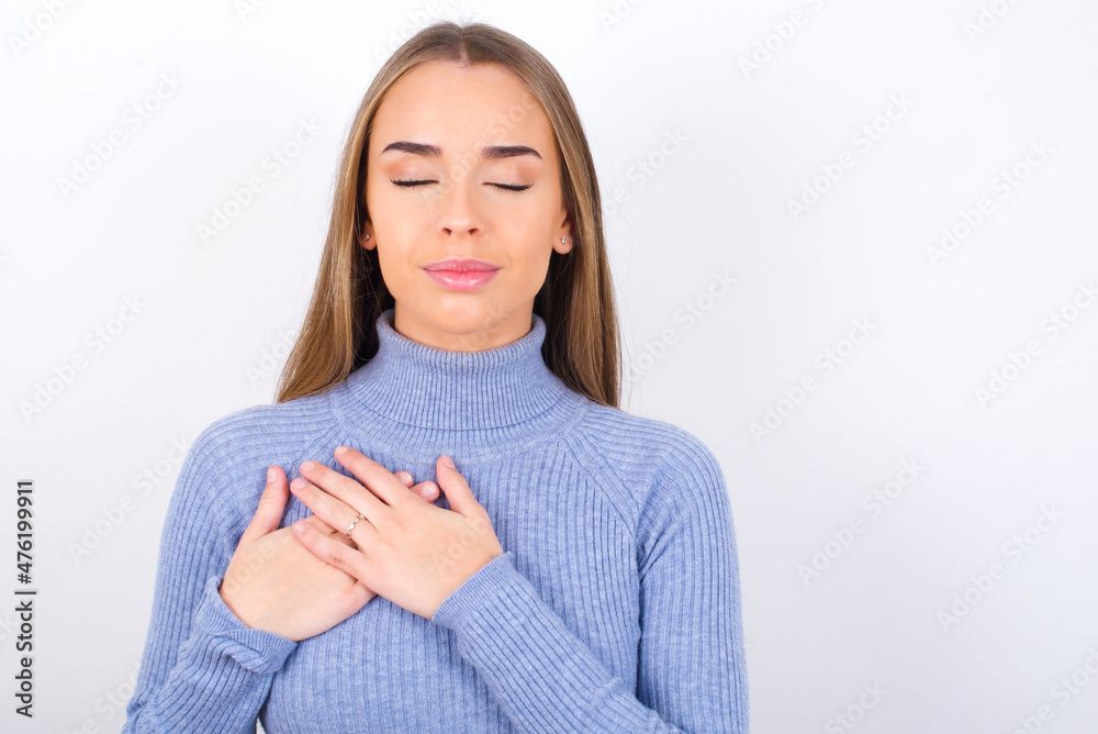 Young caucasian girl wearing blue turtleneck over whcloses eyes and keeps hands on chest near heart, expresses sincere emotions, being kind hearted and honest. Body language and real feelings concept. Young caucasian girl wearing blue turtleneck over whcloses eyes and keeps hands on chest near heart, expresses sincere emotions, being kind hearted and honest. Body language and real feelings concept.