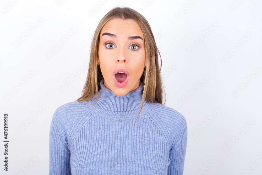 Young caucasian girl wearing blue turtleneck over white background having stunned and shocked look, with mouth open and jaw dropped exclaiming: Wow, I can't believe this. Surprise and shock Young caucasian girl wearing blue turtleneck over white background having stunned and shocked look, with mouth open and jaw dropped exclaiming: Wow, I can't believe this. Surprise and shock