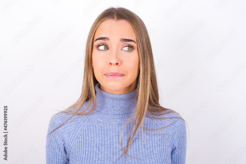 Amazed puzzled Young caucasian girl wearing blue turtleneck over white background , curves lips and has worried look, sees something awful in front. Amazed puzzled Young caucasian girl wearing blue turtleneck over white background , curves lips and has worried look, sees something awful in front.