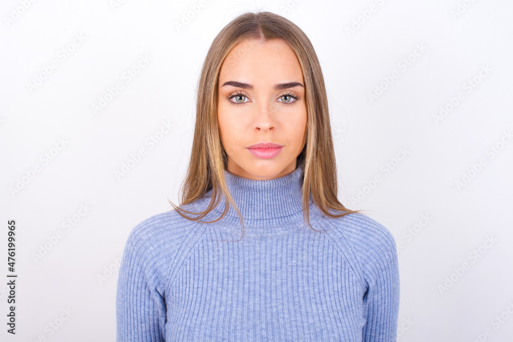 Joyful Young caucasian girl wearing blue turtleneck over white background looking to the camera, thinking about something. Both arms down, neutral facial expression. Joyful Young caucasian girl wearing blue turtleneck over white background looking to the camera, thinking about something. Both arms down, neutral facial expression.