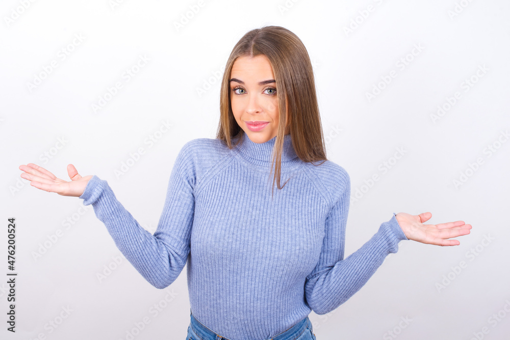 Puzzled and clueless Young caucasian girl wearing blue turtleneck over white background with arms out, shrugging shoulders, saying: who cares, so what, I don't know. Negative human emotions. Puzzled and clueless Young caucasian girl wearing blue turtleneck over white background with arms out, shrugging shoulders, saying: who cares, so what, I don't know. Negative human emotions.