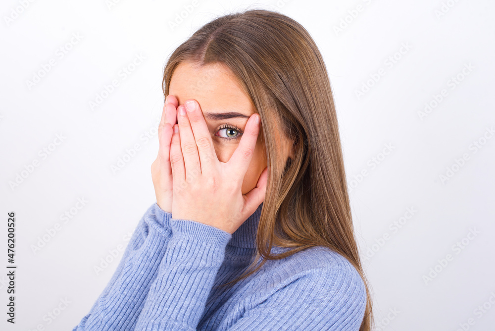 Young caucasian girl wearing blue turtleneck over white background covering face with hands and peering out with one eye between fingers. Scared from something or someone. Young caucasian girl wearing blue turtleneck over white background covering face with hands and peering out with one eye between fingers. Scared from something or someone.