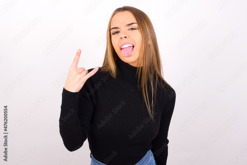 Portrait of a crazy Young caucasian girl wearing black turtleneck over white background showing tongue horns up gesture, expressing excitement of being on concert of band. Portrait of a crazy Young caucasian girl wearing black turtleneck over white background showing tongue horns up gesture, expressing excitement of being on concert of band.
