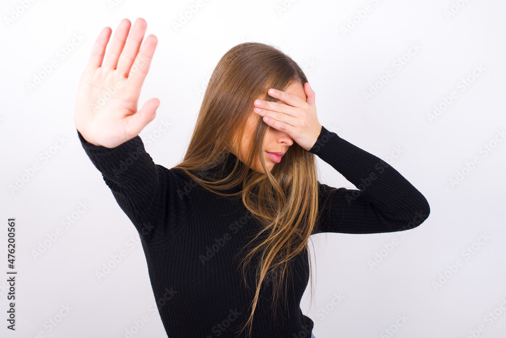 Young caucasian girl wearing black turtleneck over white background covers eyes with palm and doing stop gesture, tries to hide. Don't look at me, I don't want to see, feels ashamed or scared. Young caucasian girl wearing black turtleneck over white background covers eyes with palm and doing stop gesture, tries to hide. Don't look at me, I don't want to see, feels ashamed or scared.