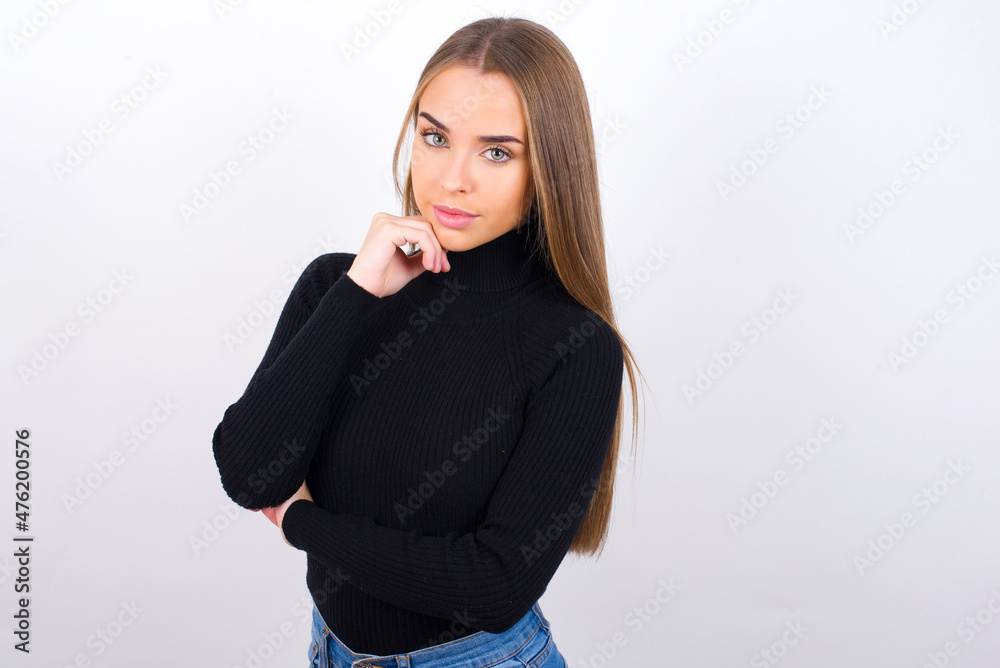 Face expressions and emotions. Thoughtful Young caucasian girl wearing black turtleneck over white background holding hand under his head, having doubtful look. Face expressions and emotions. Thoughtful Young caucasian girl wearing black turtleneck over white background holding hand under his head, having doubtful look.
