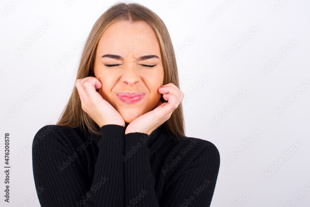 Portrait of Young caucasian girl wearing black turtleneck over white background being overwhelmed, expressing excitement and happiness with closed eyes and hands near face. Portrait of Young caucasian girl wearing black turtleneck over white background being overwhelmed, expressing excitement and happiness with closed eyes and hands near face.