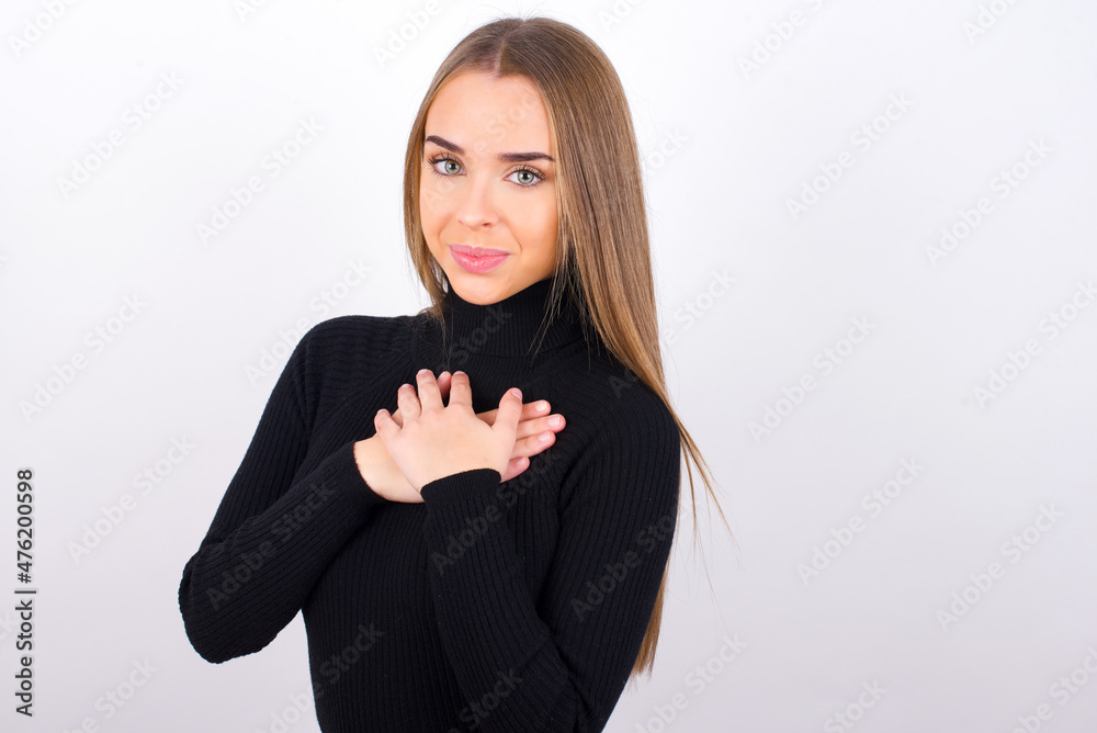 Honest Young caucasian girl wearing black turtleneck over white background keeps hands on chest, touched by compliment or makes promise, looks at camera with great pleasure. Honest Young caucasian girl wearing black turtleneck over white background keeps hands on chest, touched by compliment or makes promise, looks at camera with great pleasure.