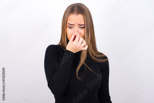 Young caucasian girl wearing black turtleneck over white background , holding his nose because of a bad smell.