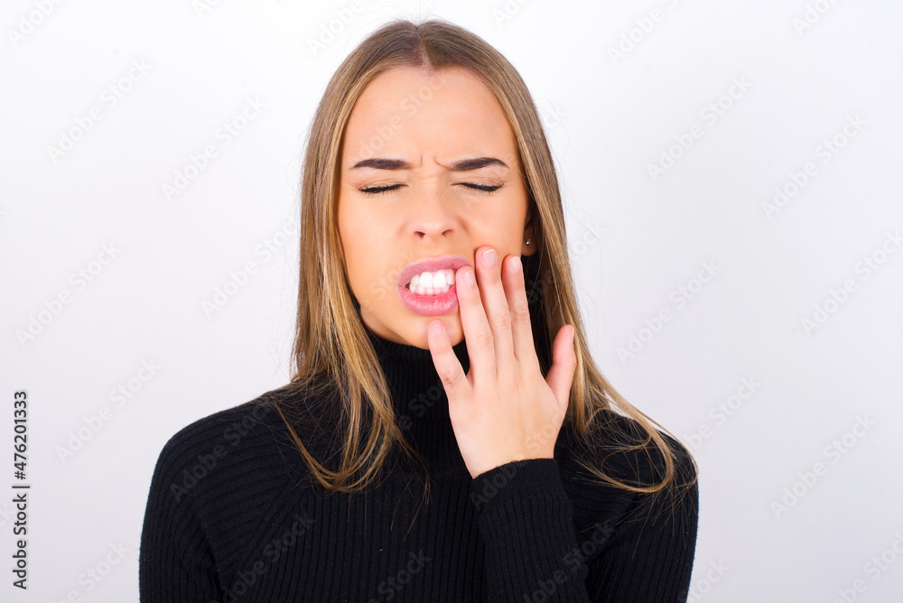 Tooth ache concept. Indoor shot of Young caucasian girl wearing black turtleneck feeling pain, holding his cheek with hand, suffering from bad toothache, looking at camera with painful expression Tooth ache concept. Indoor shot of Young caucasian girl wearing black turtleneck feeling pain, holding his cheek with hand, suffering from bad toothache, looking at camera with painful expression