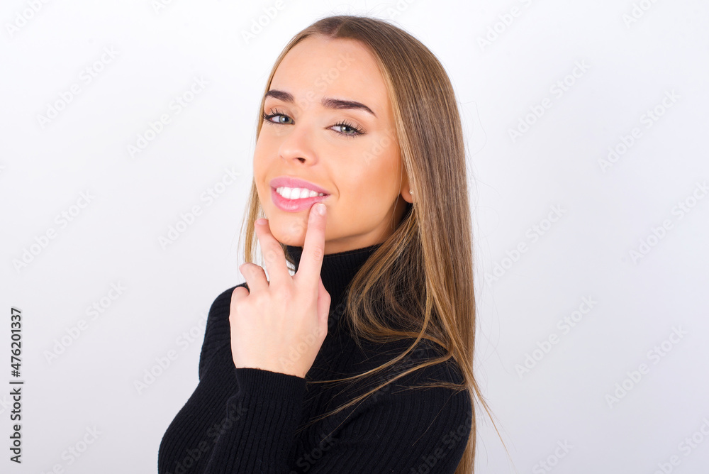 Carefree successful Young caucasian girl wearing black turtleneck over white background touching jawline gazing camera tilting head grinning white teeth delighted. Dental care concept. Carefree successful Young caucasian girl wearing black turtleneck over white background touching jawline gazing camera tilting head grinning white teeth delighted. Dental care concept.