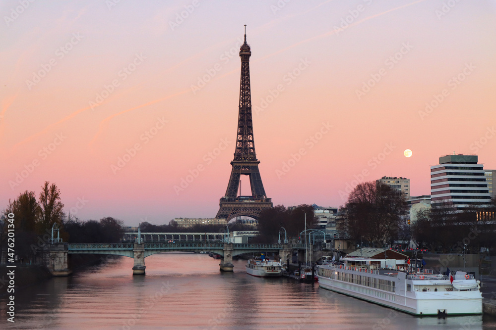 Fototapeta premium Silhouette of Eiffel tower with sunset twilight orange pink sky while full moon rising over the seine river with boats and urban modern building, trees during winter time 