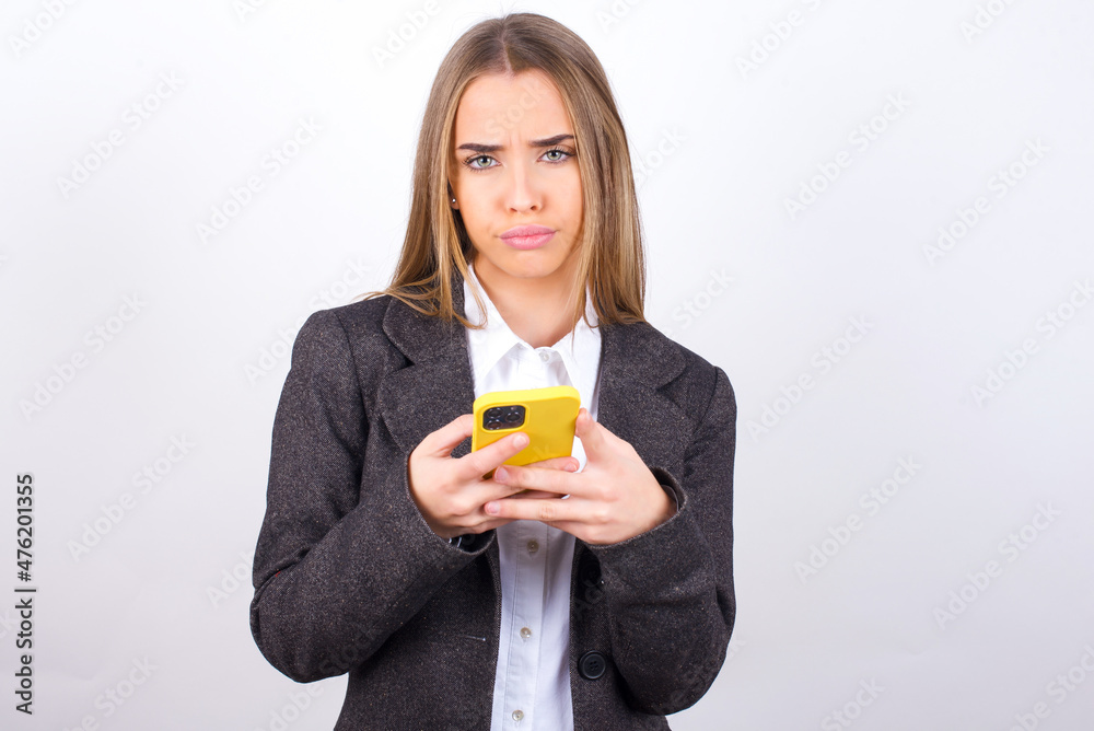 Portrait of a confused Young business woman wearing jacket over white background holding mobile phone and shrugging shoulders and frowning face. Portrait of a confused Young business woman wearing jacket over white background holding mobile phone and shrugging shoulders and frowning face.