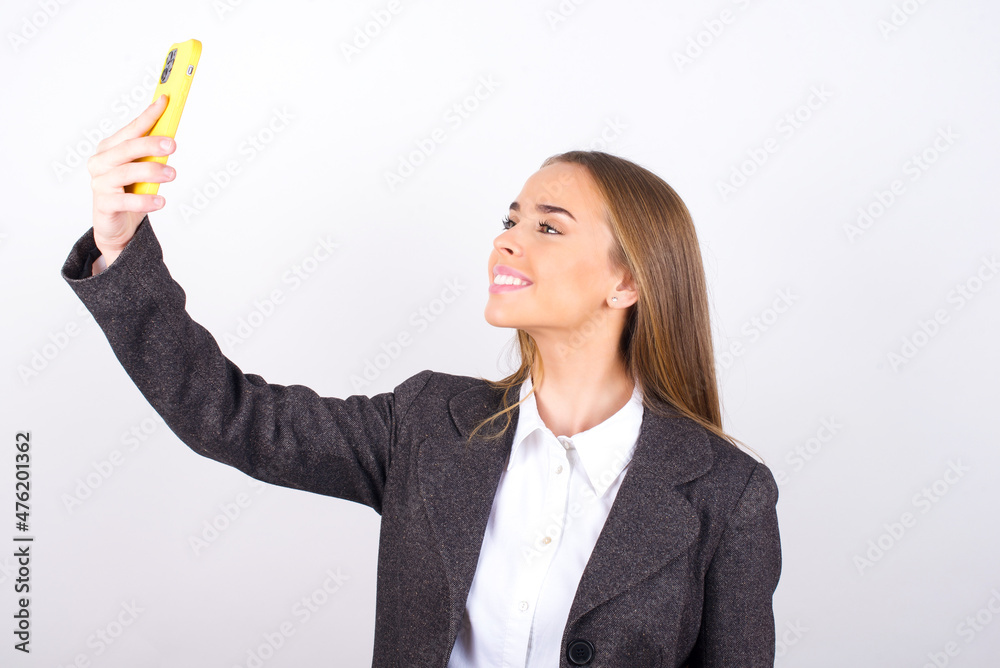 Isolated shot of pleased cheerful Young business woman wearing jacket over white background , makes selfie with mobile phone. People, technology and leisure concept Isolated shot of pleased cheerful Young business woman wearing jacket over white background , makes selfie with mobile phone. People, technology and leisure concept