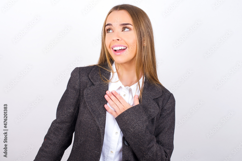 Joyful Young business woman wearing jacket over white background expresses positive emotions recalls something funny keeps hand on chest and giggles happily. Joyful Young business woman wearing jacket over white background expresses positive emotions recalls something funny keeps hand on chest and giggles happily.