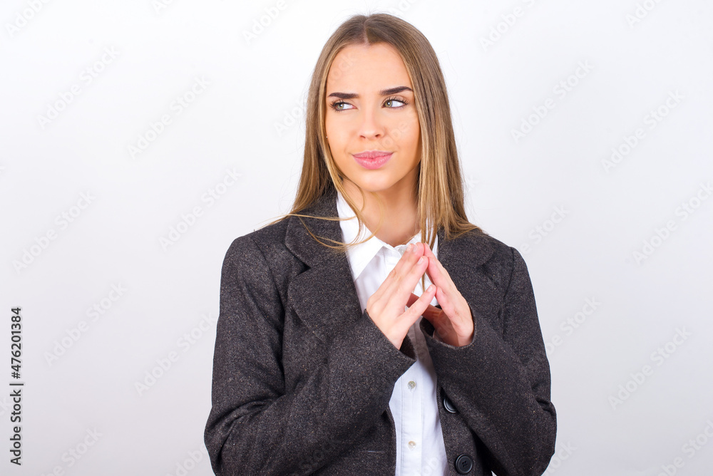 Young business woman wearing jacket over white background steepls fingers and looks mysterious aside has great evil plan in mind Young business woman wearing jacket over white background steepls fingers and looks mysterious aside has great evil plan in mind