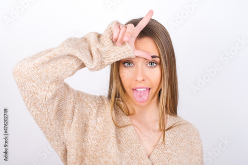 Konstfotografi Funny  young caucasian girl wearing knitted sweater over white background makes loser gesture mocking at someone sticks out tongue making grimace face