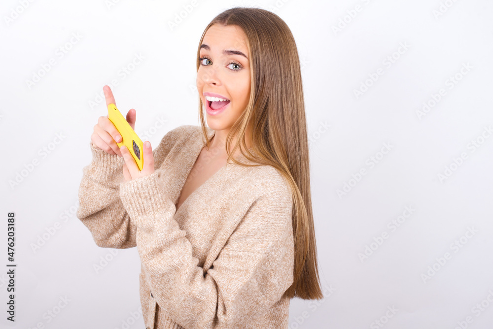 Nice addicted cheerful young caucasian girl wearing knitted sweater over white background using gadget playing network game Nice addicted cheerful young caucasian girl wearing knitted sweater over white background using gadget playing network game