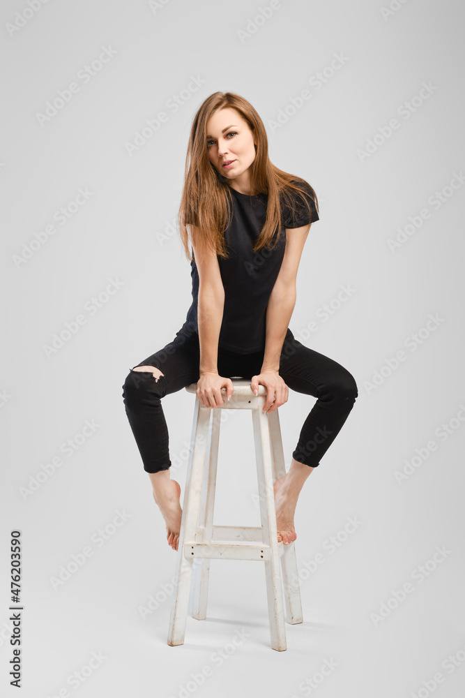 Full length studio portrait of young barefoot woman in jean and shirt sitting on a chair
