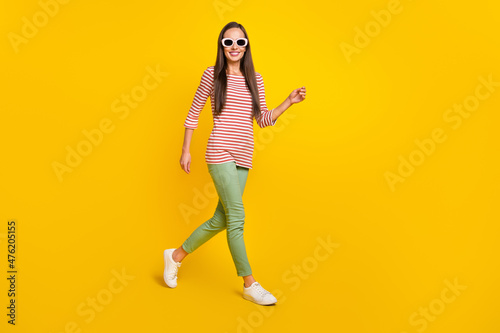 Photo of pretty shiny young woman wear striped shirt dark glasses smiling walking isolated yellow color background